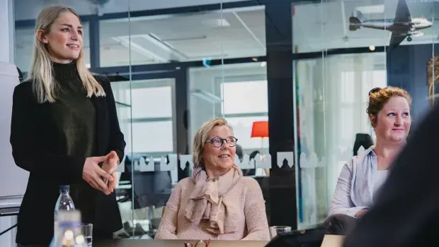 Kvinna står upp i konferensrum med fingertopparna ihopa. Två kvinnor sitter ner. Alla tittar åt höger